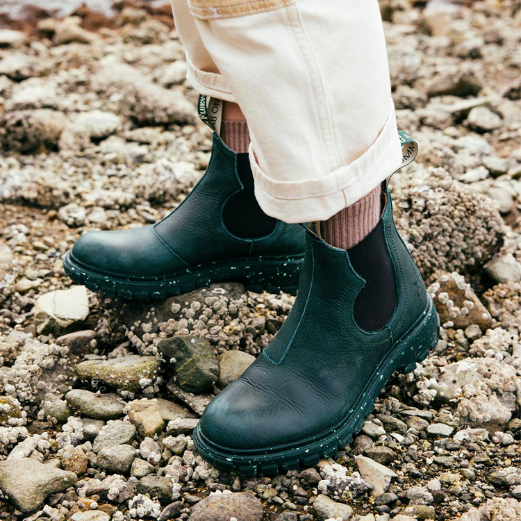 Image of a person from the ankles down wearing the Fluevog x Nimmo Bay Tursday boots on a pebble beach terrain.