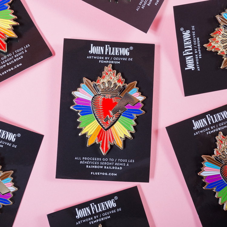 Several Fluevog Pride Pins scattered over a pink background. Each pin is attached to a black card stock package with text: "John Fluevog / artwork by / oevre de Femporium / All proceeds go to / Tous les bénéfices seront remis à / Rainbow Railroad / fluevog.com"