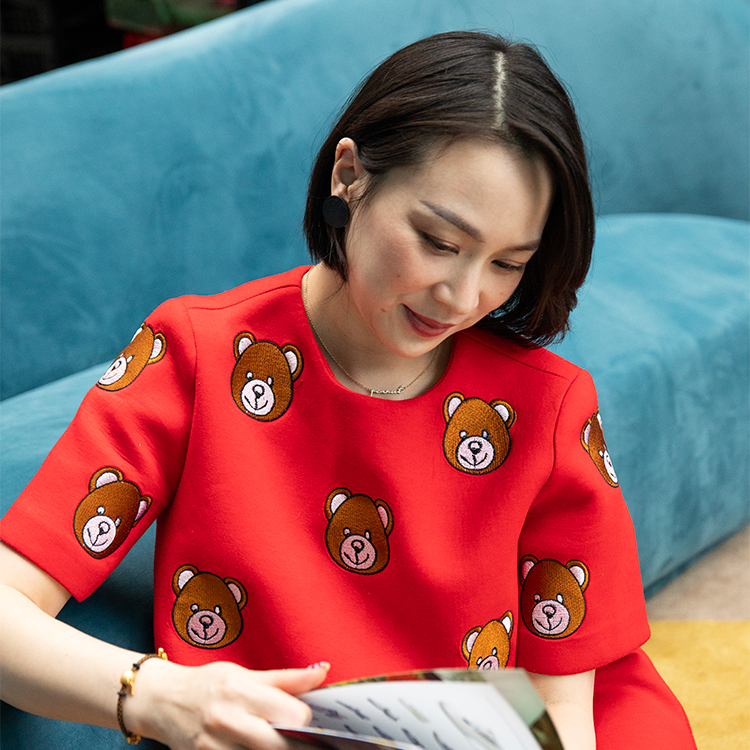 A woman wears a red dress decorated with teddy bears while reading a book and leaning against a blue couch.