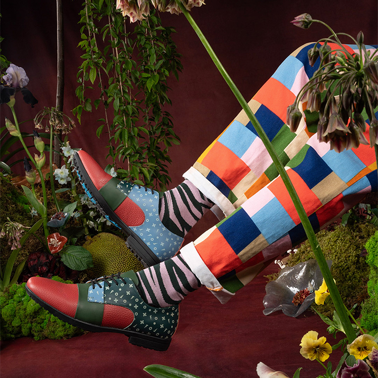 A model wears the Prophesy Fuller lace-up shoes, the green/pink Sargent Vog Socks, and a pair of pants with brightly-coloured repeating squares.
